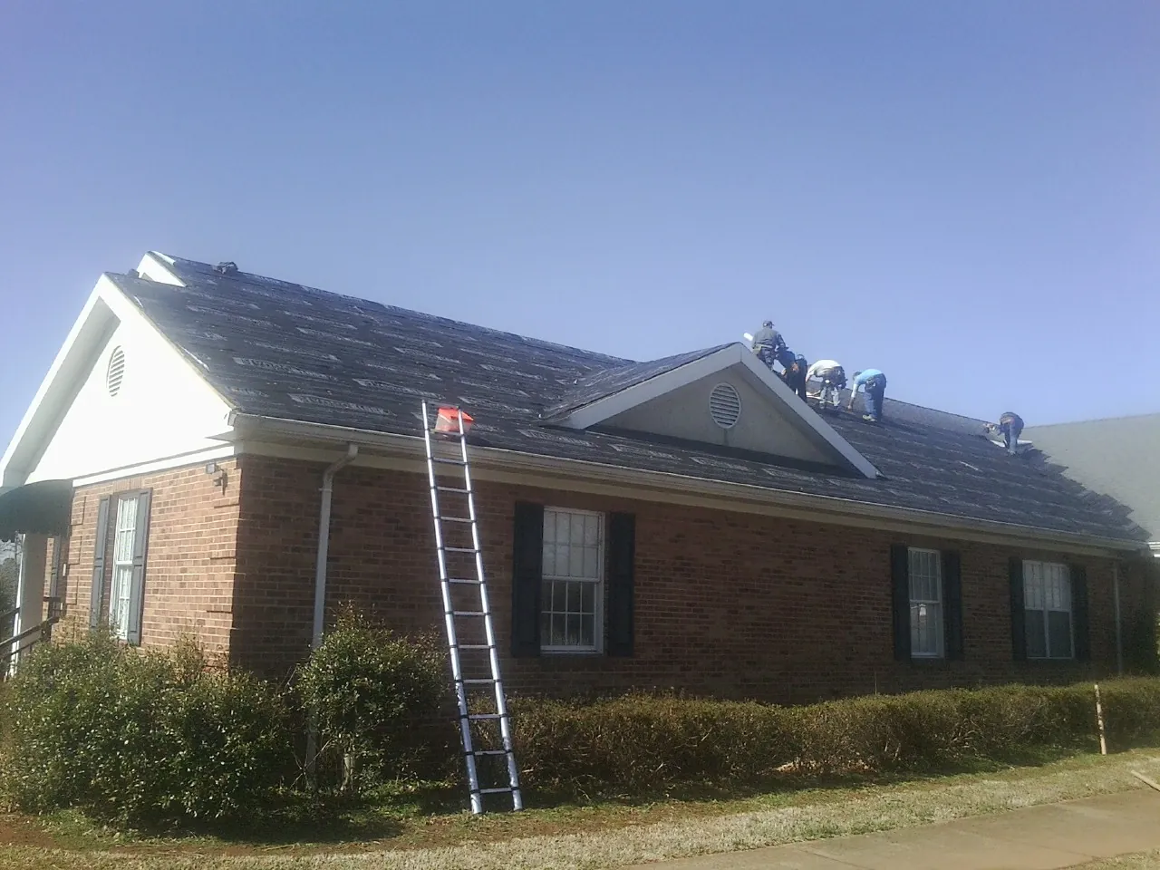 Crew tearing shingles off a brick ranch roof with a ladder set — Metal Roof Repair work by our crew in Hopkinton