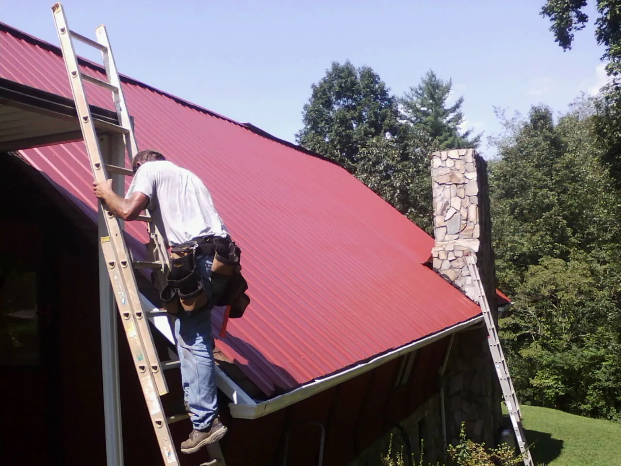 Roofer on a ladder beside a red metal roof and stone chimney — Soffit & Fascia Repair by Roofing Brothers in Hopkinton